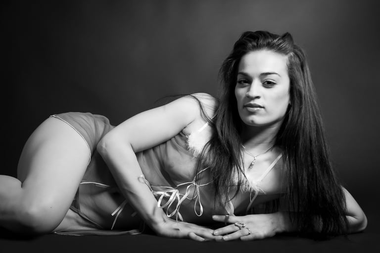 Femme allongée sur le côté, portant une nuisette transparente avec des détails en plumes, posant sur un fond noir dans une ambiance boudoir en noir et blanc.