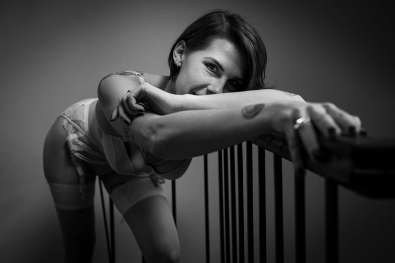 Femme appuyée sur une rampe d escalier en lingerie blanche et bas, son sourire espiègle et ses tatouages ajoutant du caractère à cette image boudoir saisissante.