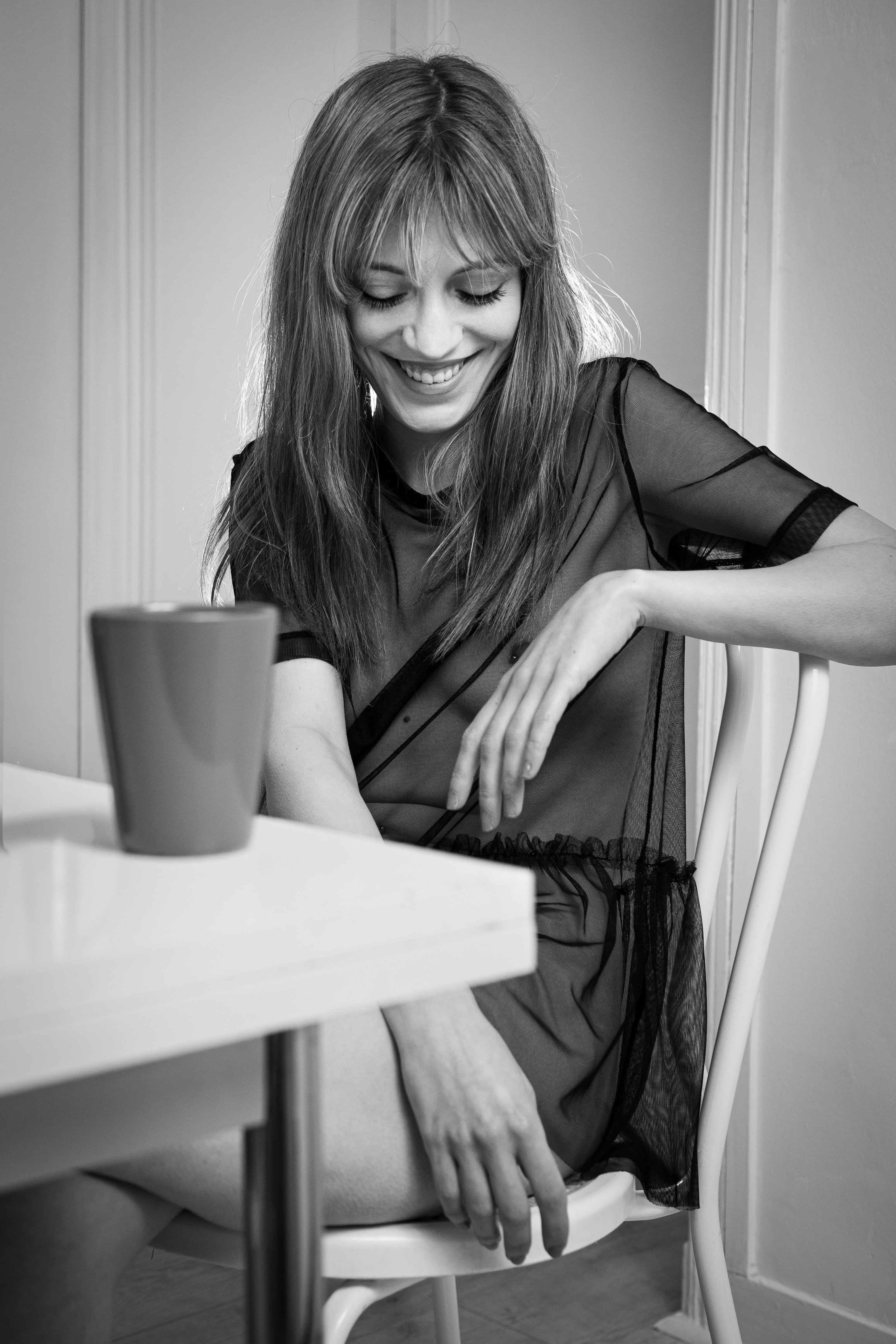 Femme souriante assise à une table blanche, vêtue d'une tenue noire transparente, capturée en noir et blanc doux pour une ambiance boudoir détendue et naturelle.