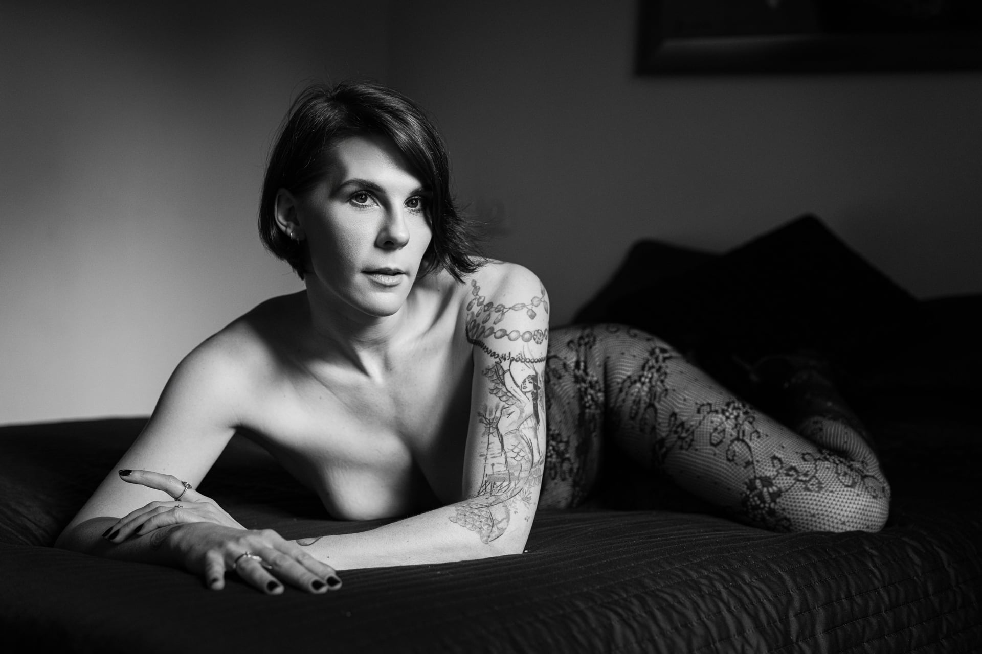 Femme allongée sur un lit en collants en dentelle, ses tatouages et son regard intense créant un portrait boudoir captivant et intime.