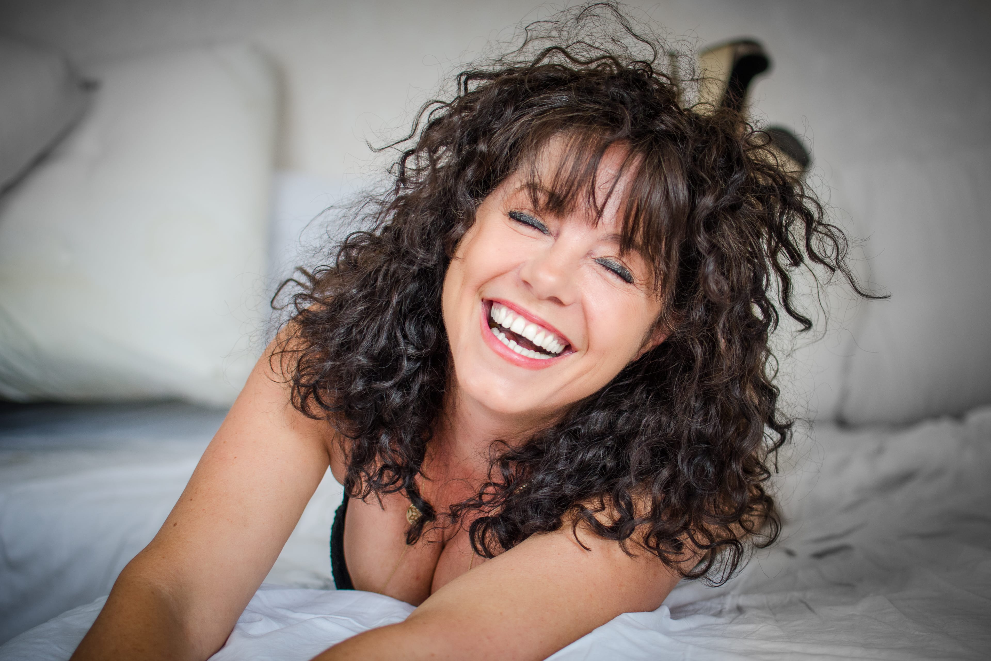 Femme joyeuse allongée sur un lit blanc, ses cheveux bouclés encadrant son visage, capturée dans un moment boudoir lumineux et ludique de bonheur insouciant.